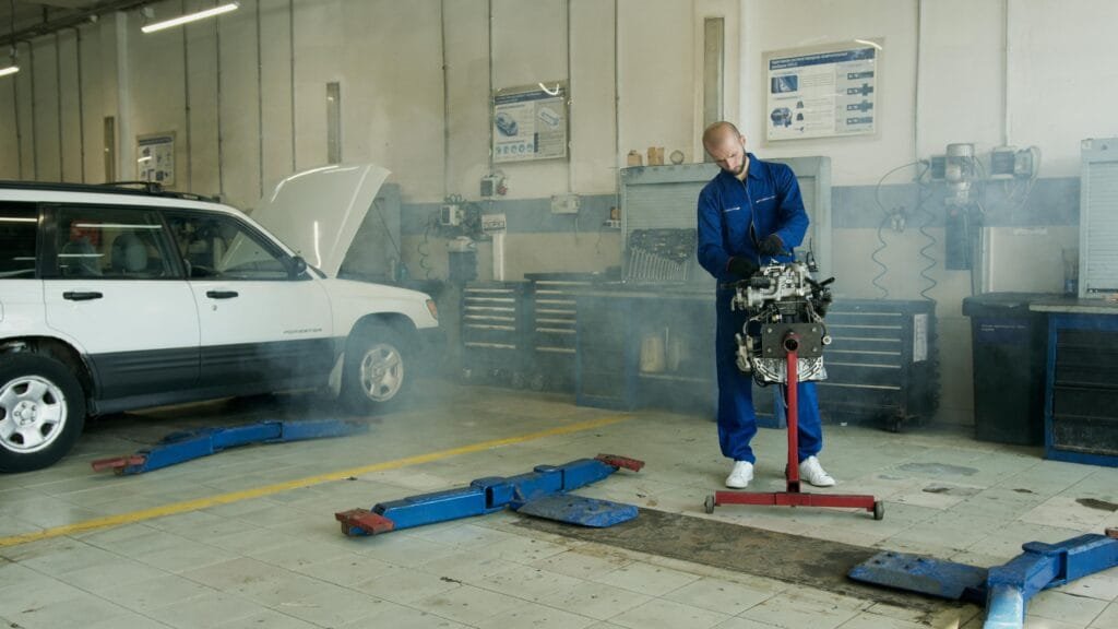 Mechanic in blue overalls working on a car engine in an auto repair shop with a white car