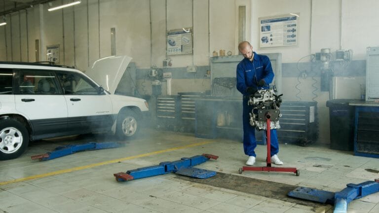 Mechanic in blue overalls working on a car engine in an auto repair shop with a white car